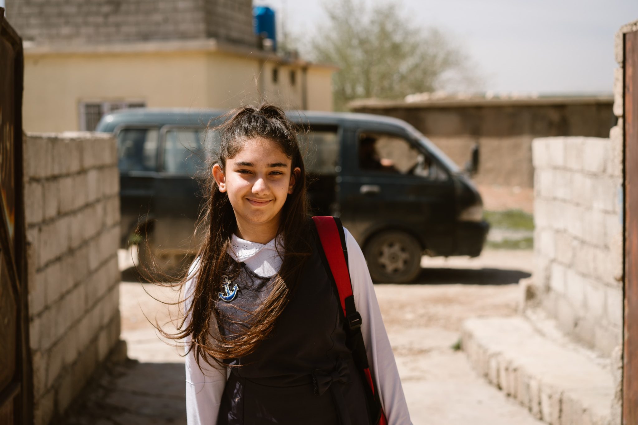 Tumma pitkähiuksinen koulutyttö hymyilee koulupuvussaan asuinalueen pihamaalla.