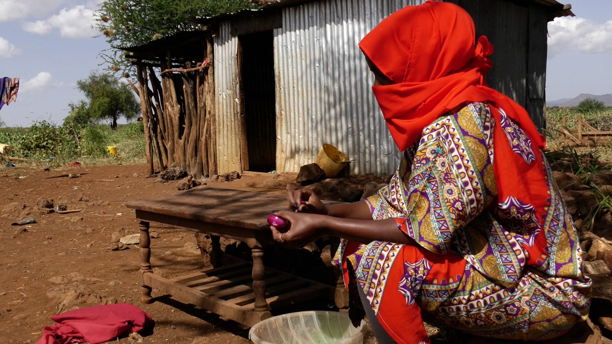 Punaiseen huiviin kietoutunut nainen istuu peltikappaleista kyhätyn rakennuksen edessä pöydän ääressä Itä-Afrikassa.