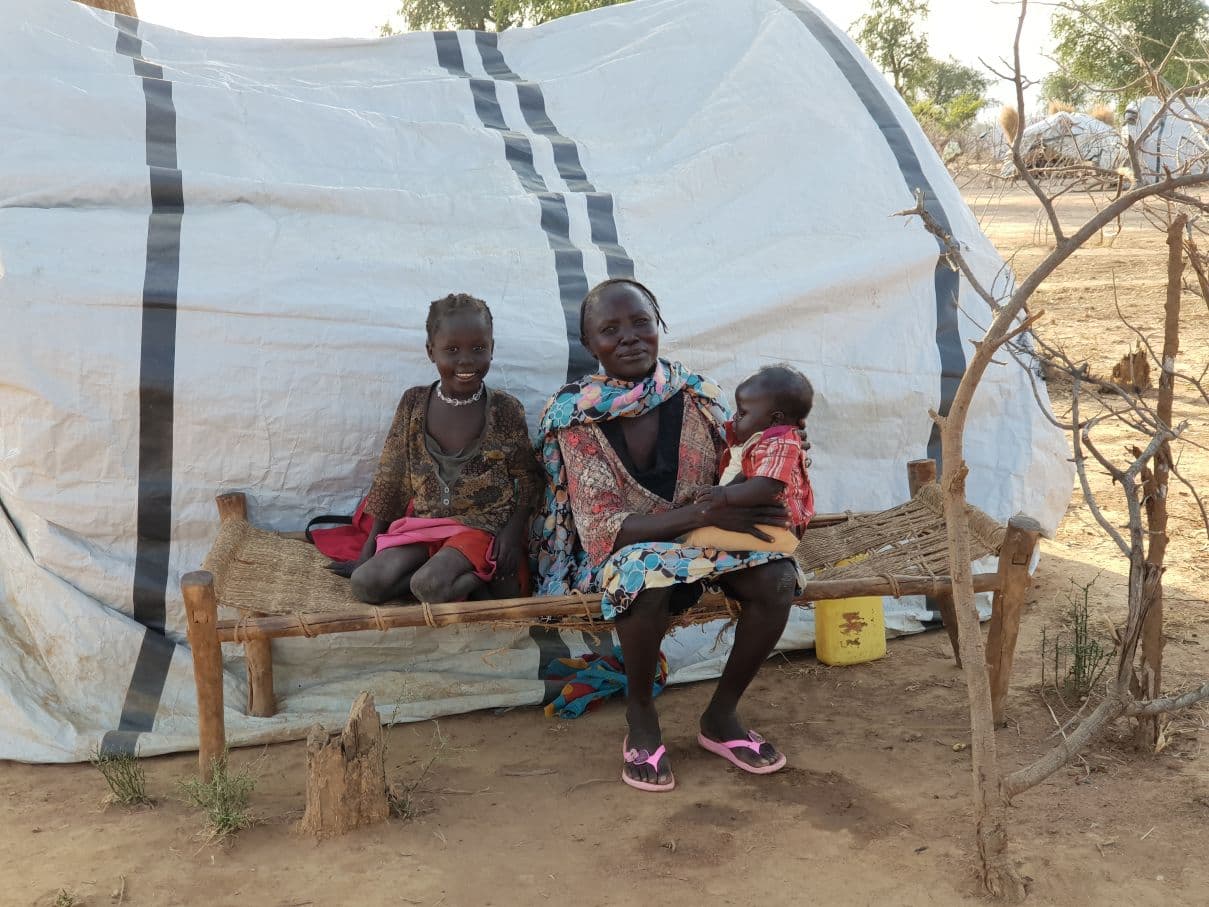 Sudanilainen nainen istuu penkillä tilapäisen teltan edustalla kahden pienen lapsen kanssa.