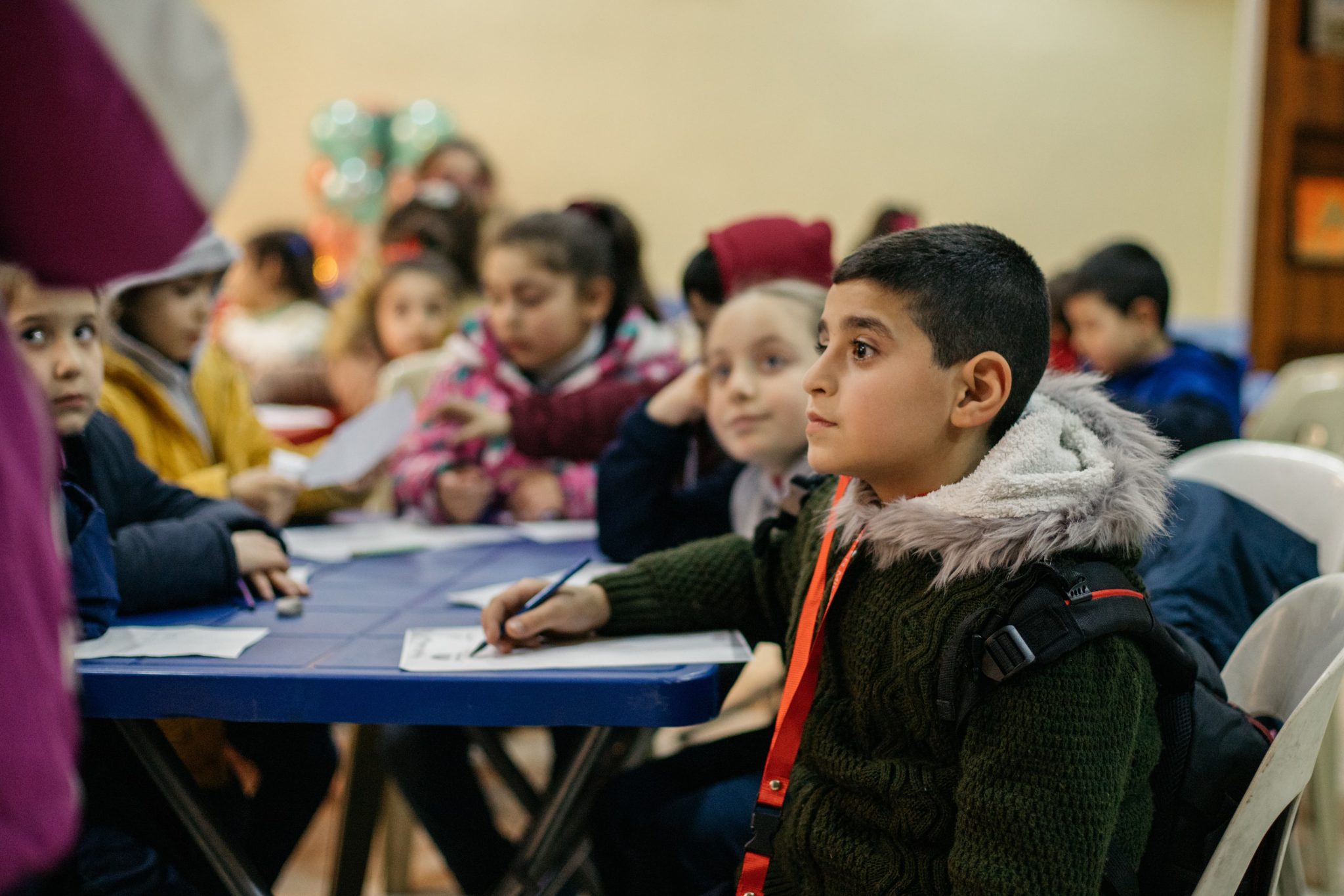 Syyrialaispoika istuu mietteliäänä koululuokassa muiden lasten keskellä ja kuuntelee opettajaa.