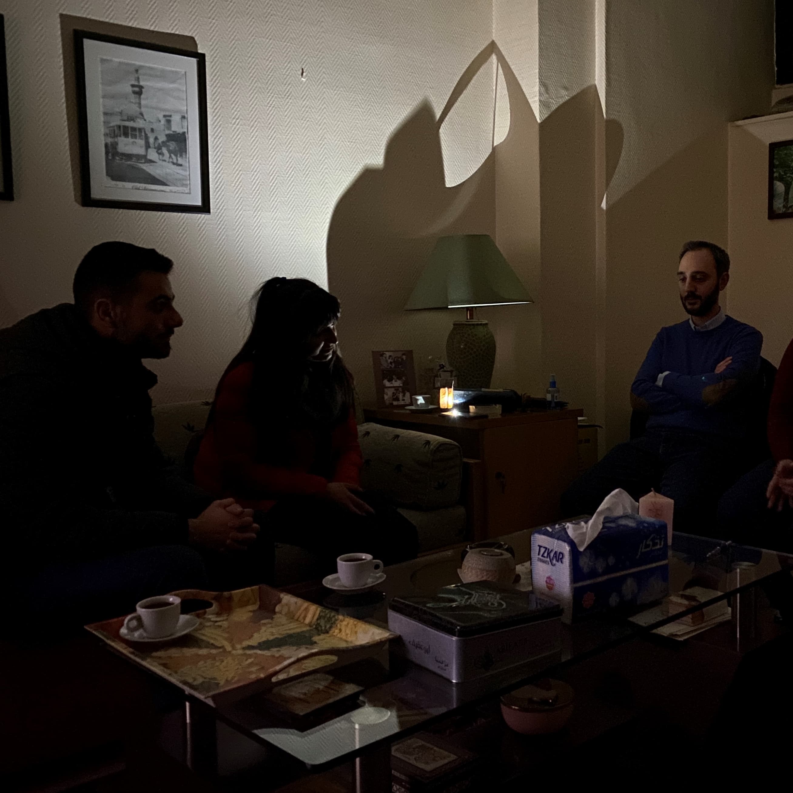Kolme syyrialaista istuu hämärässä huoneessa kännykän valossa pöydän äärellä.