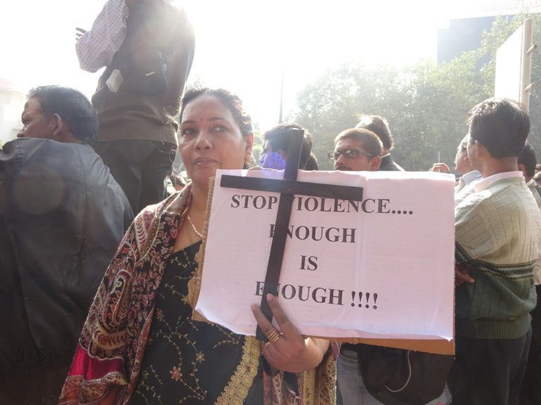 Nainen pitelee ristiä sekä lappua, jossa lukee "Stop violence.. Enough is enough!". Taustalla ihmisiä.