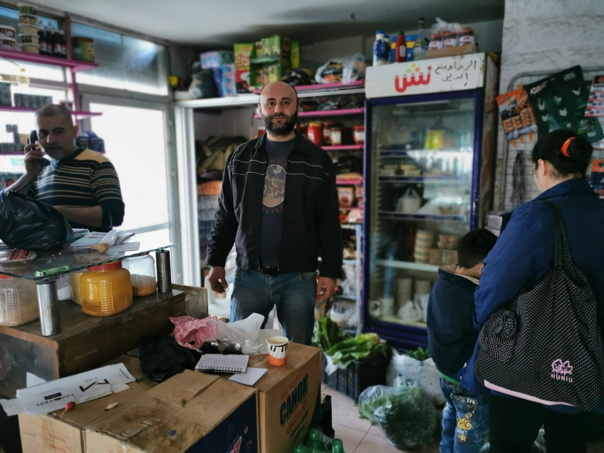 Ihmisiä syyrialaisessa kaupassa. Edustalla tiski ja pahvilaatikoita, taustalla kaupan hyllyjä ja jääkaappi, lattialla koreissa vihanneksia.
