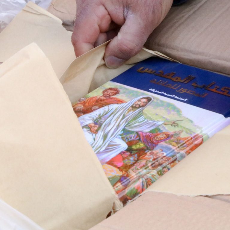 Käsi, joka siirtää pakkauspaperia syrjään kuvitetun Raamatun päältä pahvilaatikossa.