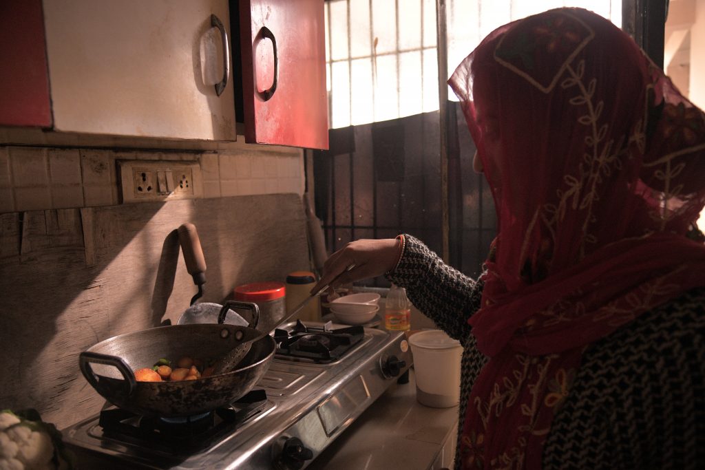 Kuvassa on Mehr, jolla on päässä punainen läpikuultava huntu. Hän on keittiossä tekemässä ruokaa paistinpannulla, joka on metallisen keittolevytason päällä. 