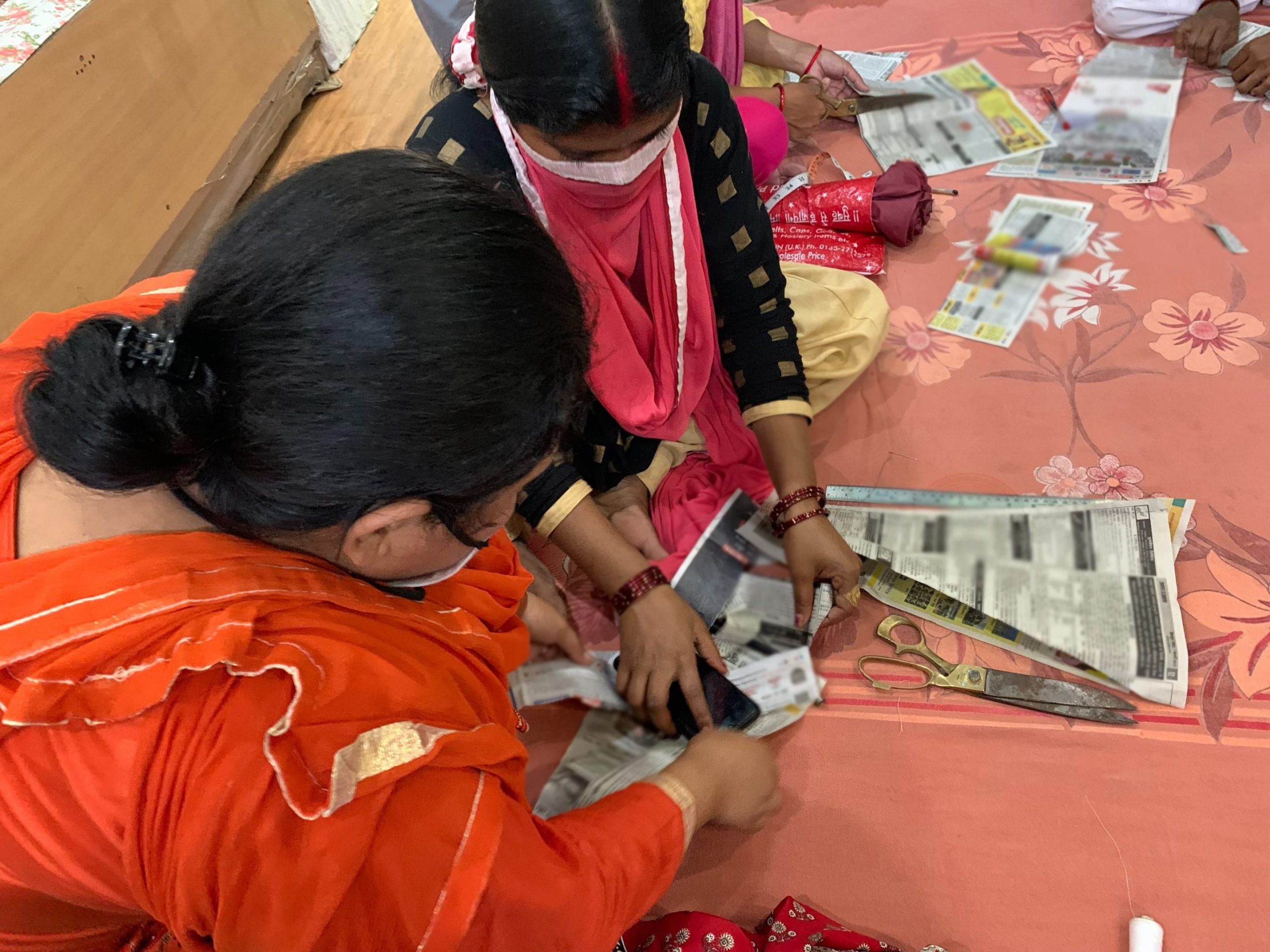 Kaksi naista istuu lattialla leikkelemässä sanomalehden kaltaista paperia. Toisella naisella on oranssinpunainen asu, toisella keltainen mekko, musta paita ja pinkki huivi.