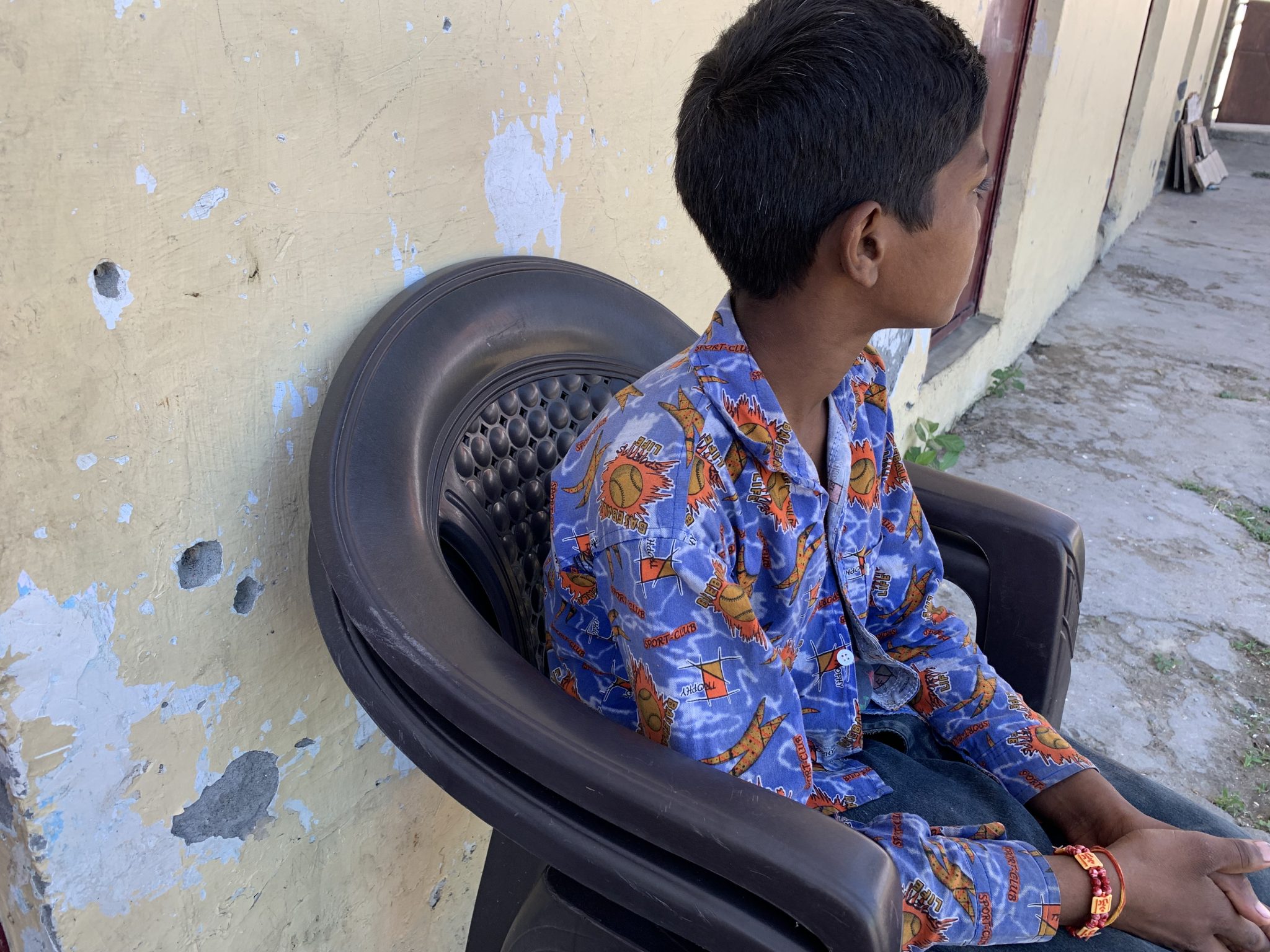 Geetikan poika istuu ulkona talon seinän vierustalla mustalla muovituolilla. Hänen kasvonsa ovat pois päin kamerasta. Hänellä on mustat hiukset ja kirjava sini-oranssi -kauluspaita päällään.