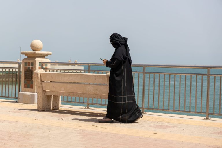 Mustiin pukeutunut nainen kävellee rantakadulla veden äärellä älypuhelin kädessä.
