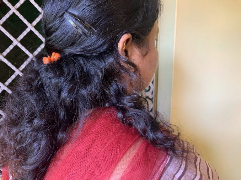 Kuvassa näkyy Shivanin pää, kasvot poispäin kamerasta. Hänellä on mustat kiharat hiukset ja punainen asu. Kuvassa näkyy oikea korva, jonka ylärustossa on punainen korvarengas.