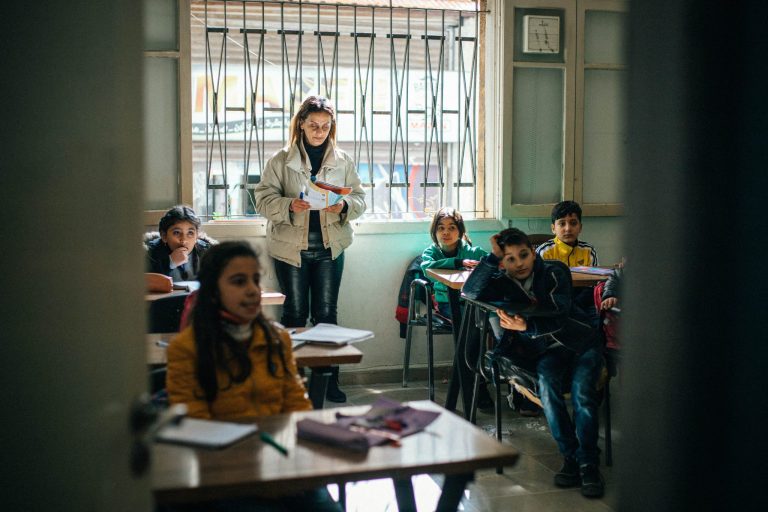 Syyrialainen opettaja Ferial seisoo luokkansa takaosassa. Luokassa on alakouluikäisiä oppilaita pulpeteissaan.