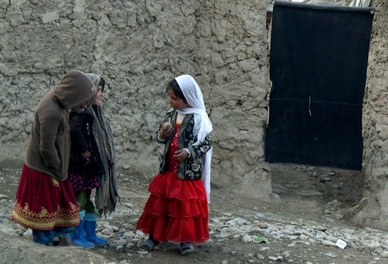 Kolme pientä lasta leikkimässä vaatimattoman kivirakennuksen edessä.