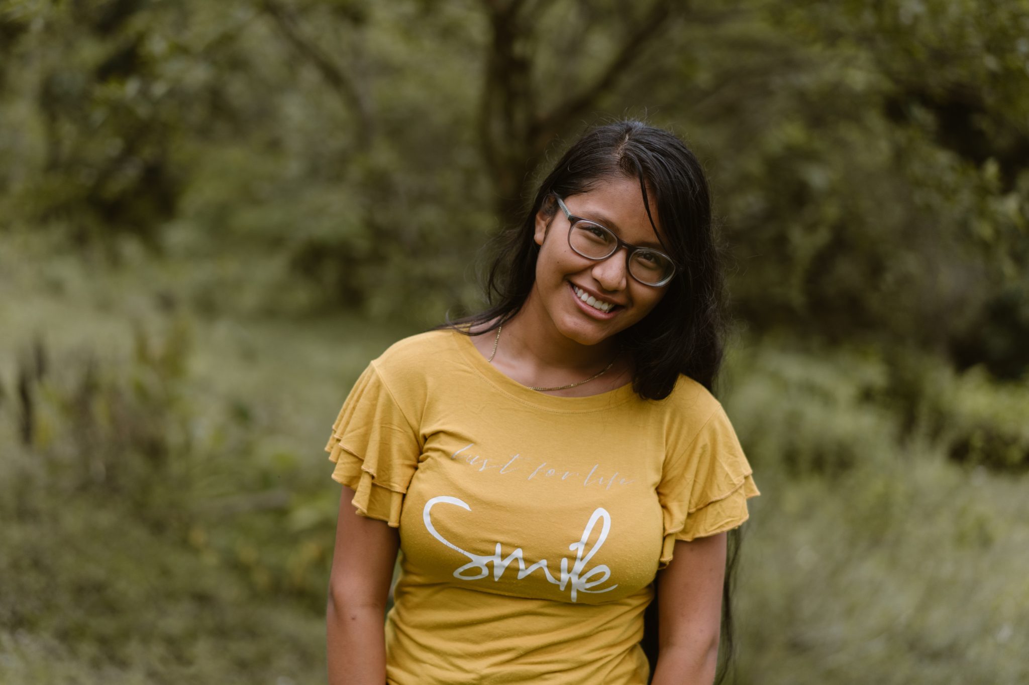 Tummatukkainen teinityttö keltaisessa paidassa hymyilee ja katsoo kameraan ulkona trooppista metsätaustaa vasten.