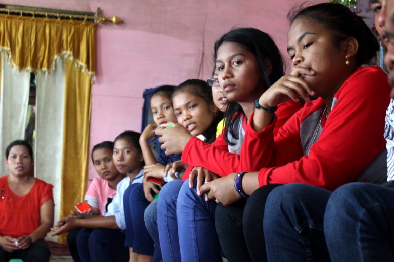 Tummatukkaiset alakouluikäiset tytöt istuvat vinosti vierekkäin ja rivin päässä näkyy heidän naispuolinen pyhäkouluopettajansa. Huoneen seinät ovat vaaleanpunaiset ja verhot keltaiset.