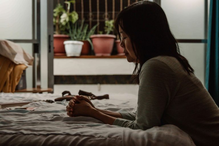 Tummahiuksinen nainen rukoilee vaaleaa sänkyä vasten polvistuneena hämärässä aasialaisessa huoneessa.