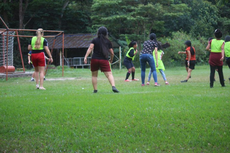 Ryhmä lasten keskuksen lapsia pelaa jalkapalloa vihreällä nurmella selkäpäin kameraan.
