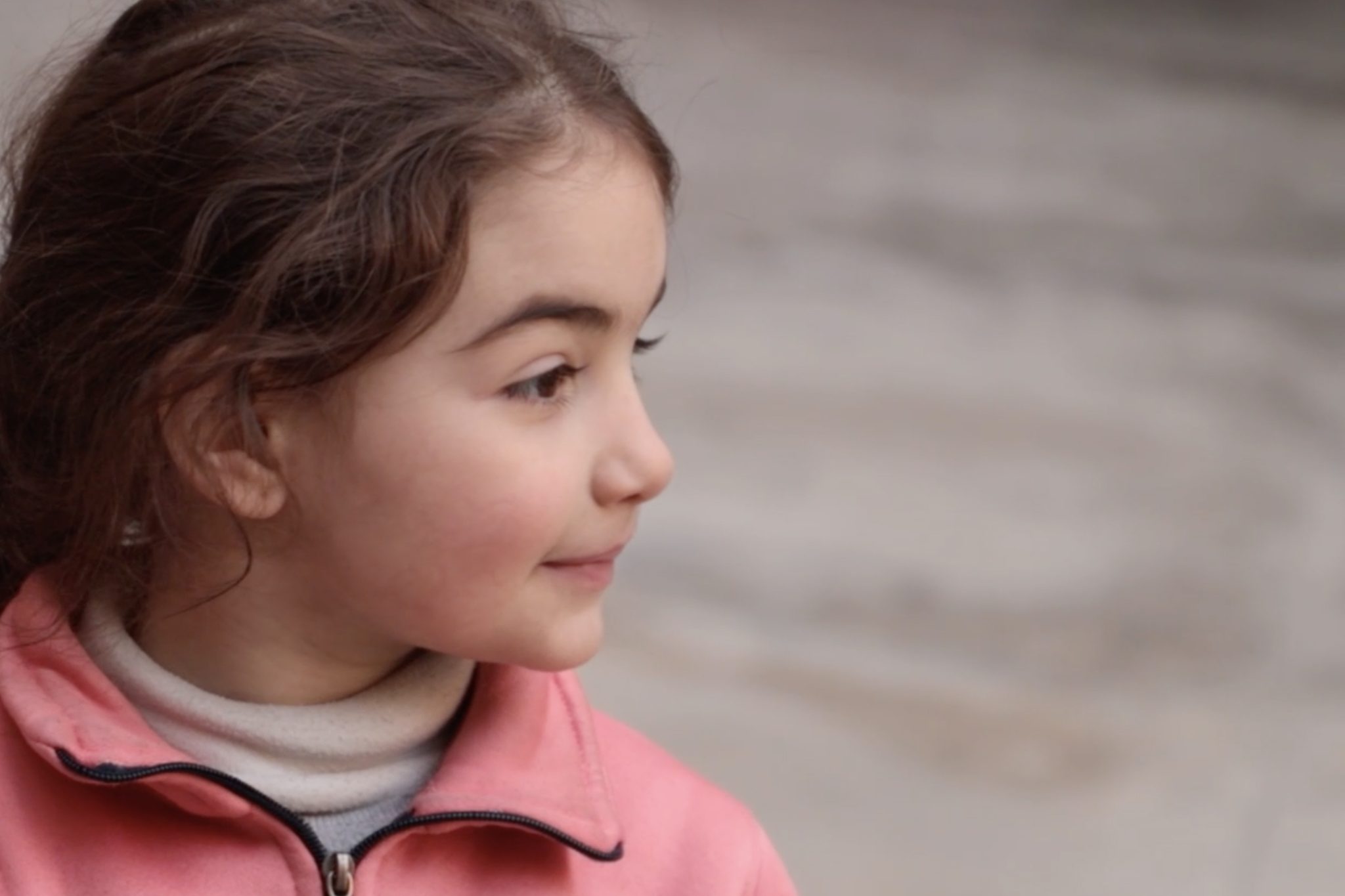 Ruskeatukkainen pikkutyttö katsoo oikealle ja hymyilee vaaleanpunaisessa takissaan.