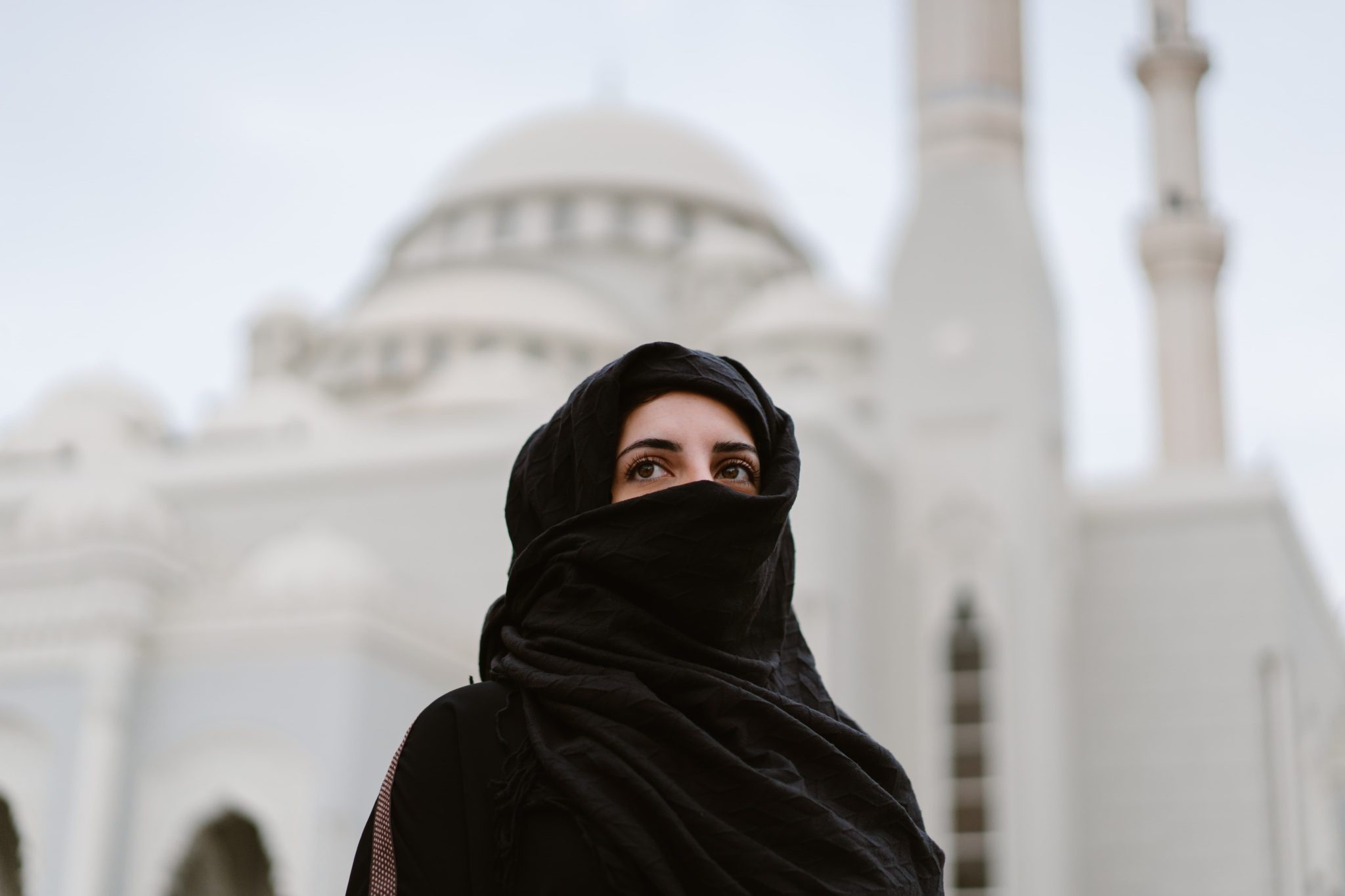 Mustaan huntuun ja kaapuun pukeutunut nainen vaalean moskeijan edessä.