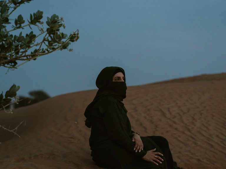 Nadia istuu aavikon hiekalla oliivipuu etualalla.