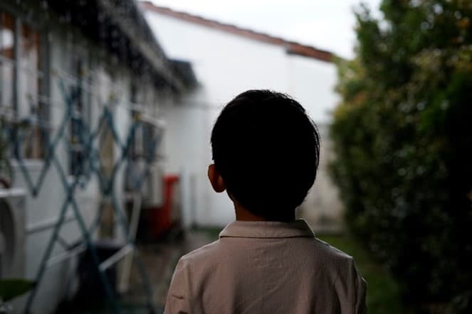 Mustatukkainen pikkupoika valkoisessa paidassa selkäpäin talon vieressä