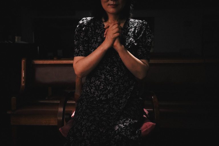 Nainen tummassa mekossa rukoilemassa pimeässä tilassa.