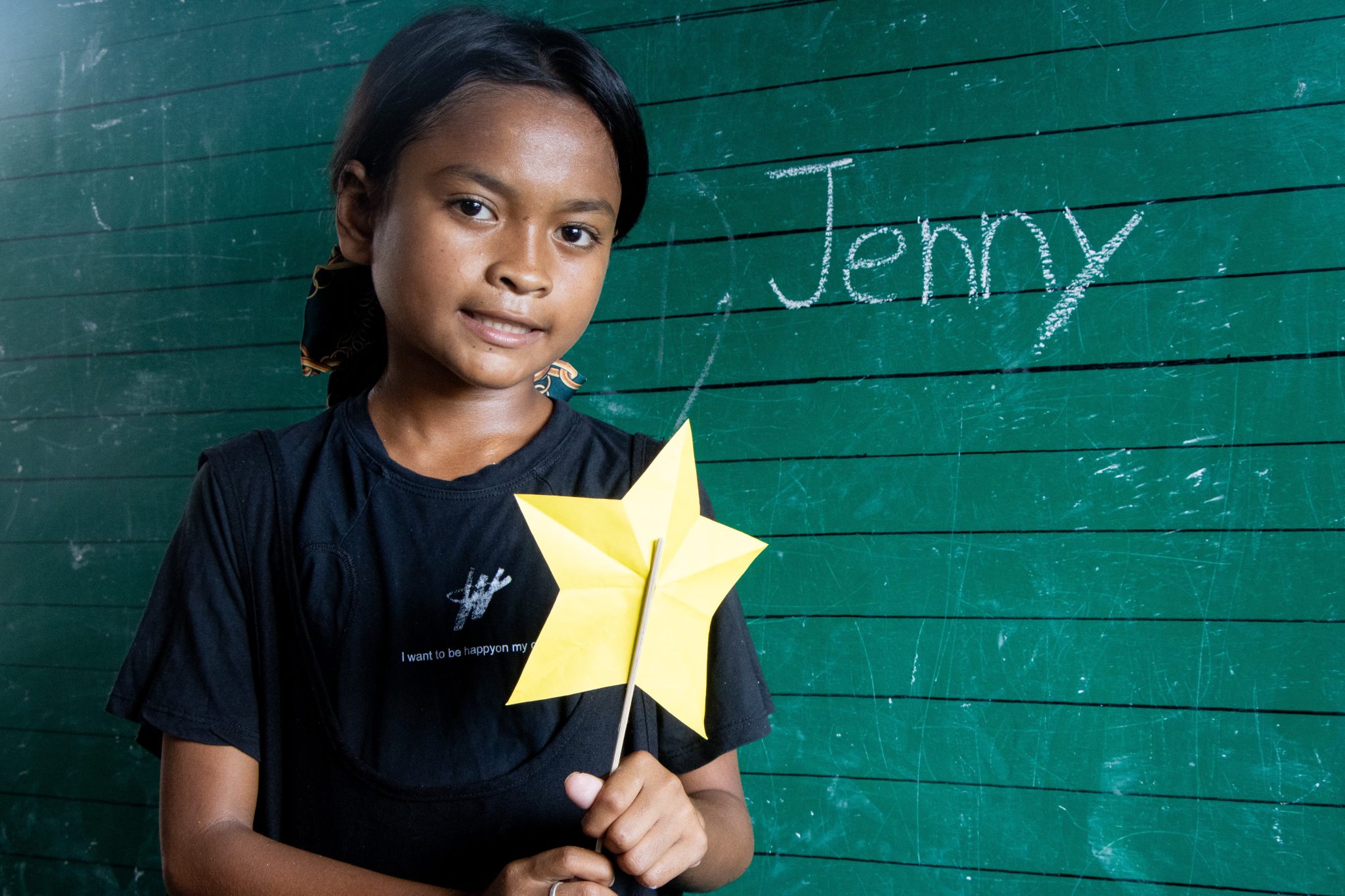 Filippiiniläinen Jenny hymyilee kameralle vihreän liitutaulun edessä. Hän pitelee askarreltua tähteä käsissään. Liitutaululla lukee hänen nimensä