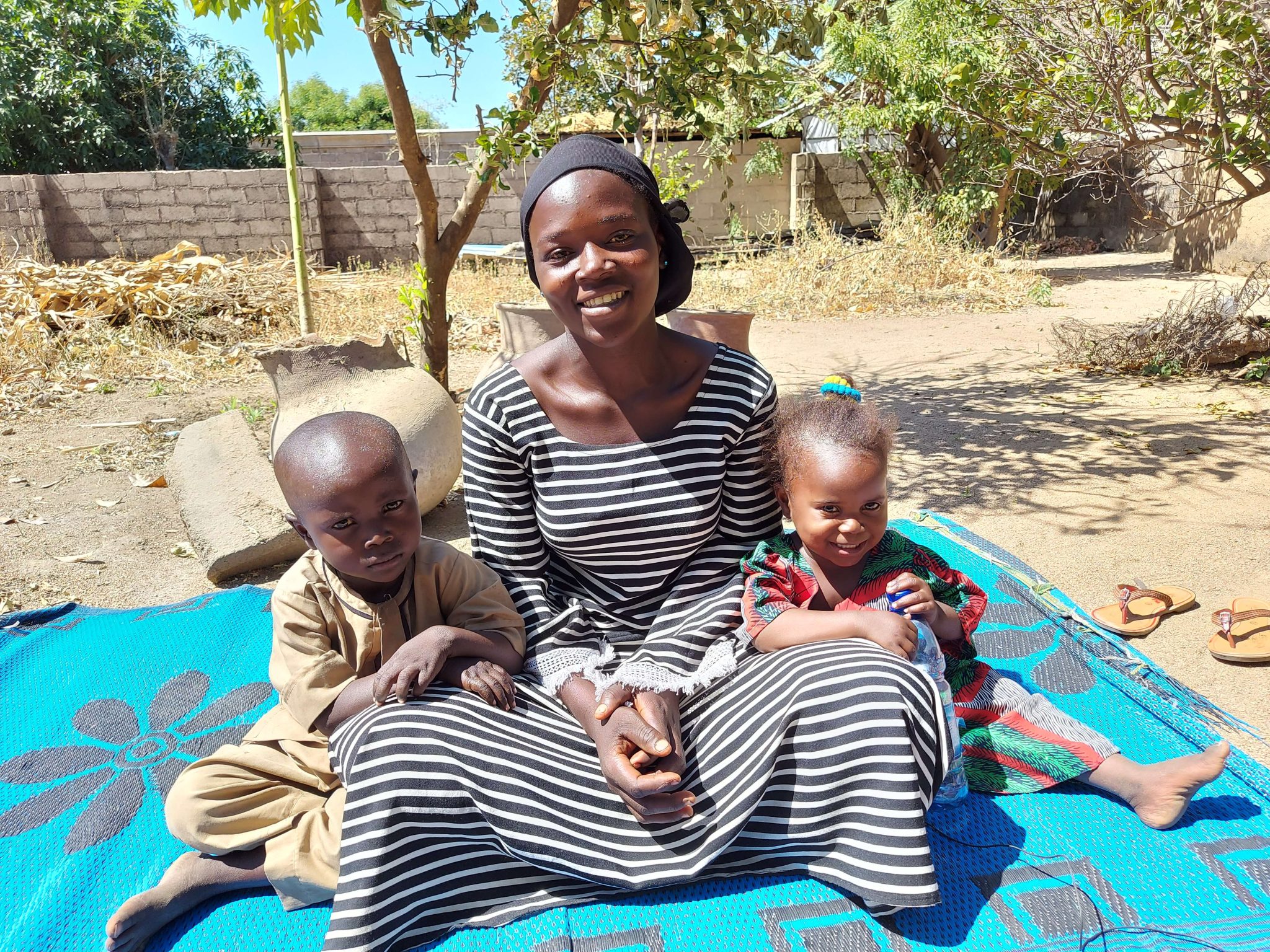 Hymyilevä nainen istuu sinisellä viltillä kahden pikkulapsen kanssa.