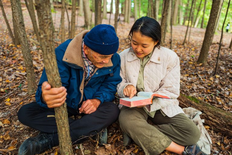 Nuori nainen istuu metsässä vanhan miehen vieressä ja he lukevat yhdessä raamattua.