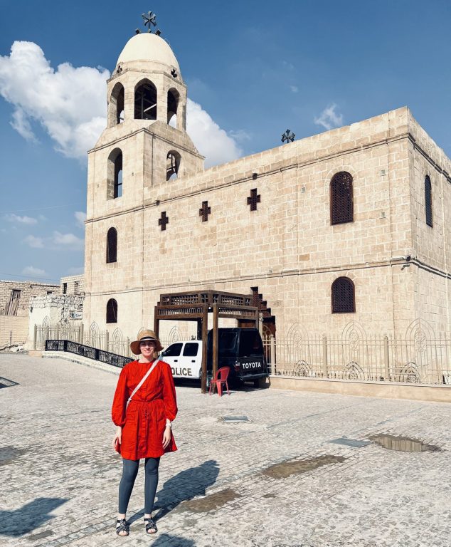 Kuvassa artikkelin kirjoittaja, Anu, seisoo Egyptiläisen beigen kirkkorakennuksen edessä. Taustalla kirkon edessä näkyy pysäköity poliisiauto.