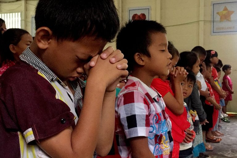 Alakouluikäisiä lapsia rivissä rukoilemassa seurakuntatilaisuudessa