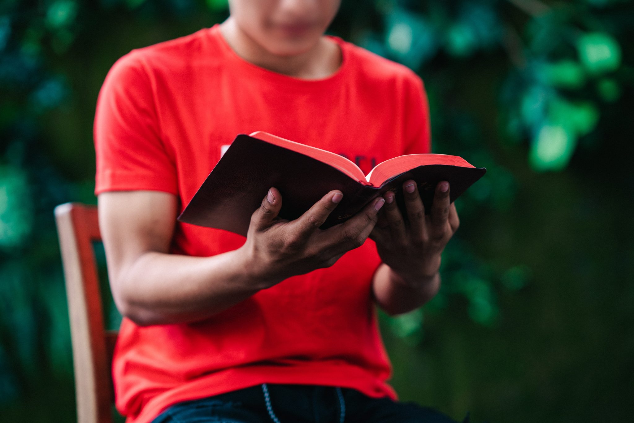 Nuori mies istuu ulkona ja lukee Raamattua. Hänen Raamattunsa on musta, punaisella reunuksella. Hänellä on yllään punainen t-paita.