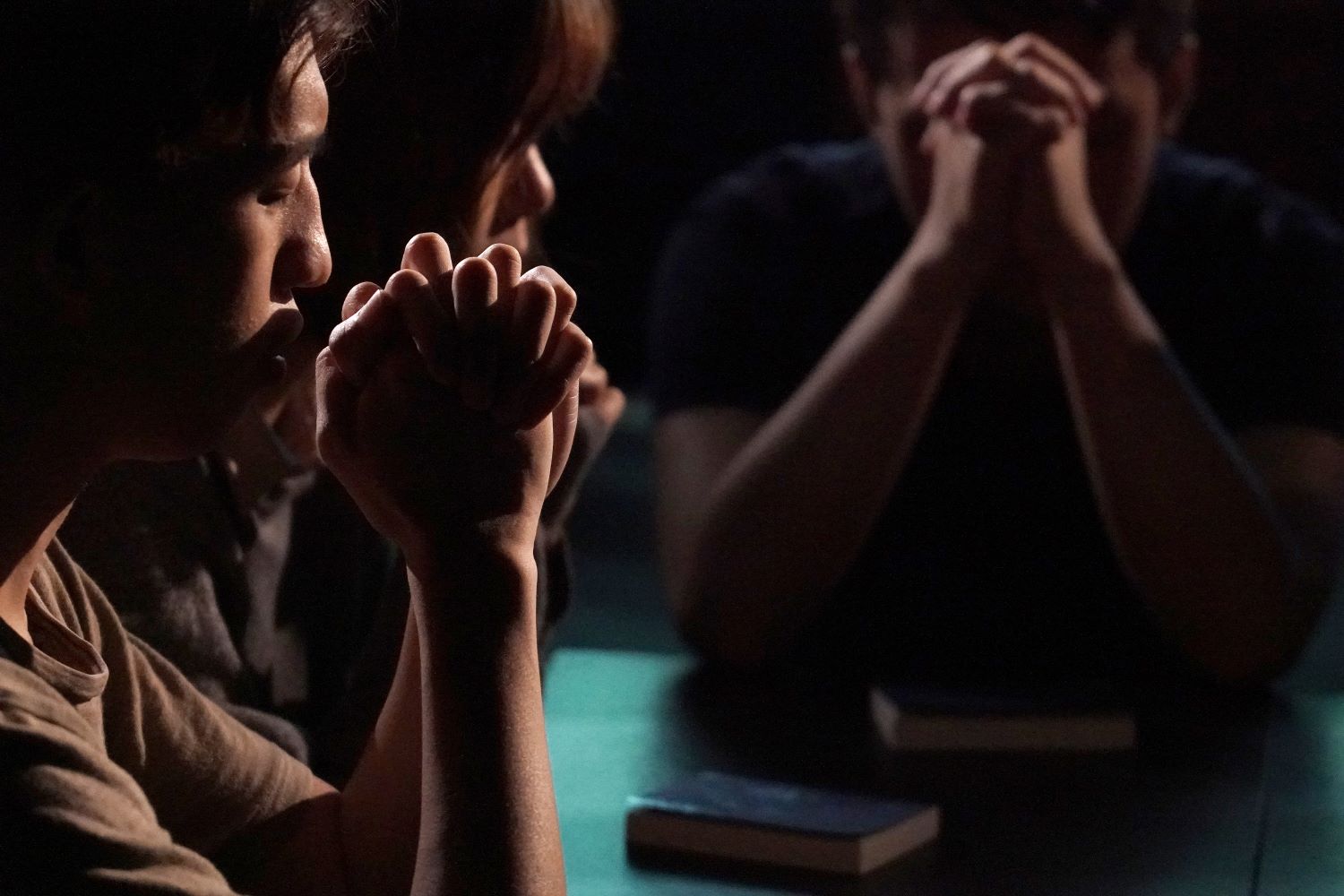 Hämärässä pöydän ääressä kolme henkilöä, jotka rukoilevat. Heidän kätensä ovat ristissä heidän kasvojensa edessä. Pöydällä kaksi kirjaa.