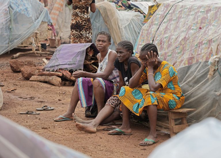 Kolme naista istuu penkillä telttojen keskellä pakolaisleirillä