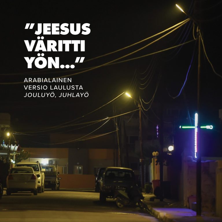 Valokuva pimeältä kadulta, jonka varrella neonvärein hohtava risti. Kuvan päällä sanat Jouluyö, juhlayö -laulun arabialaisesta versiosta: "Jeesus väritti yön".