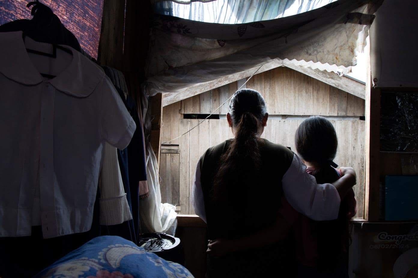 Nainen ja tyttö hämärässä talossa katsovat yhdessä pois päin kamerasta, ikkunasta ulos. Nainen on kietonut käsivartensa tytön hartioille.