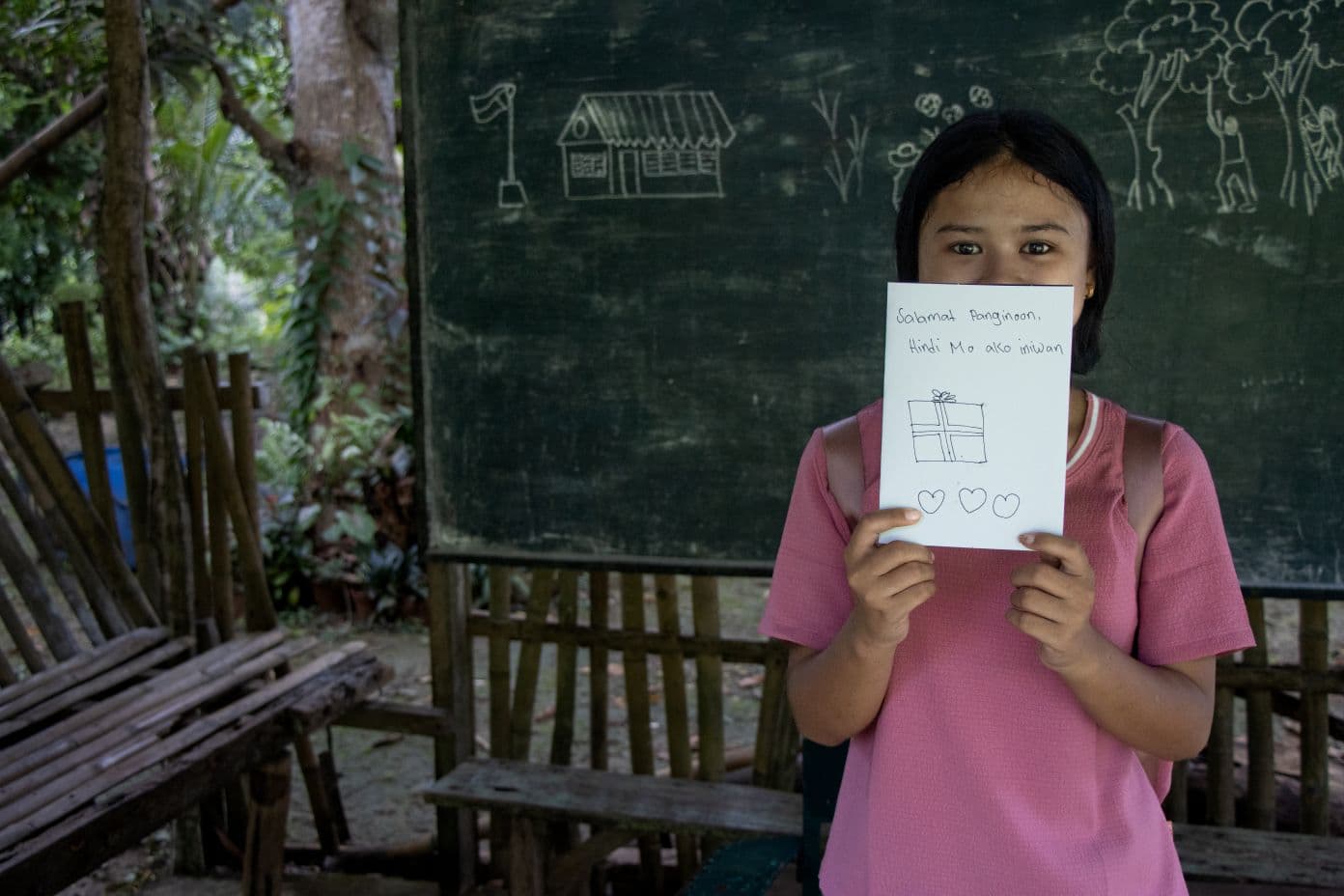 Tyttö seisoo liitutaulun edessä ja pitää kasvojensa edessä paperia, jossa on pari riviä käsin kirjoitettua tekstiä sekä piirroskuva lahjapaketista ja kolme sydäntä.