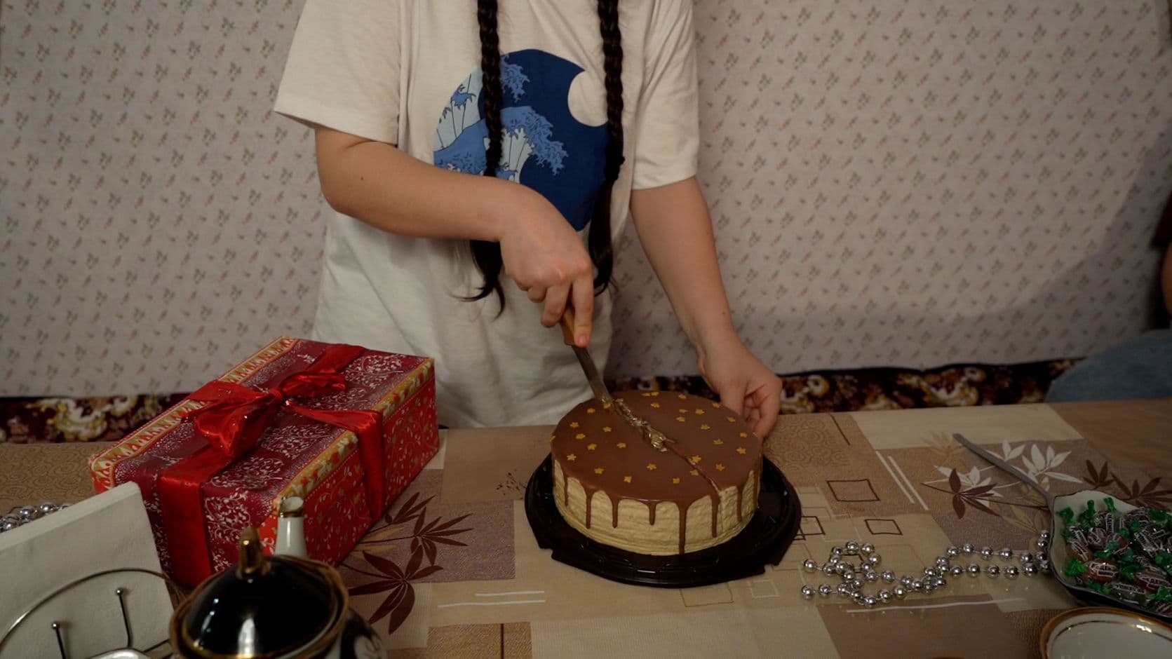 Lettipäinen tyttö leikkaa kakkua, pöydällä myös lahjapaketti.