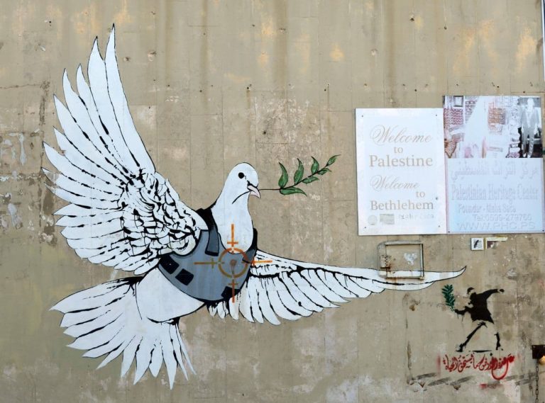 Muuriin piirretty graffiti kyyhkystä, jolla on yllään luotiliivi ja lehvä nokassaan.