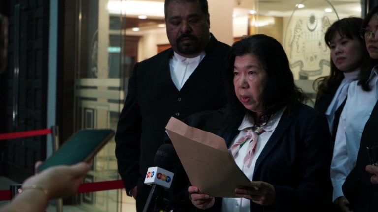 Nainen, jonka edessä on mikrofoneja. Taustalla mies ja muita ihmisiä.