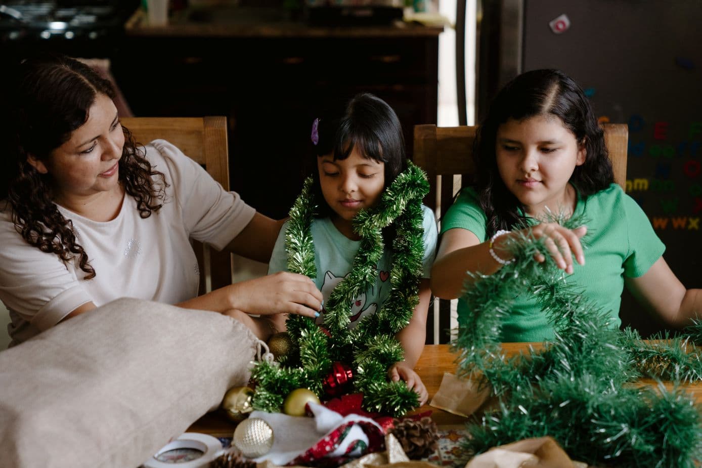 Nainen ja kaksi tyttöä istuvat pöydän ääressä, johon on levitelty joulukoristeita.