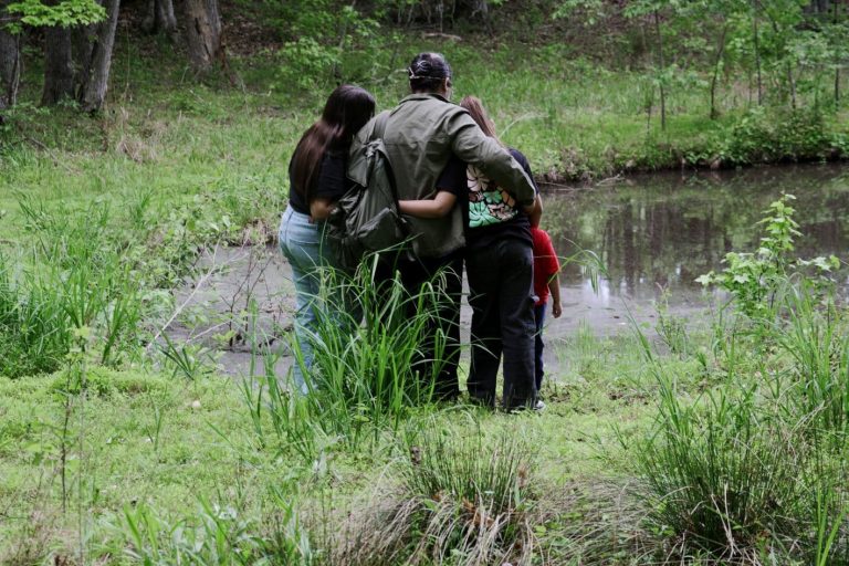 Mies ja kolme lasta selin kameraan lammen rannassa, kädet kiedottuina toistensa kauloille.