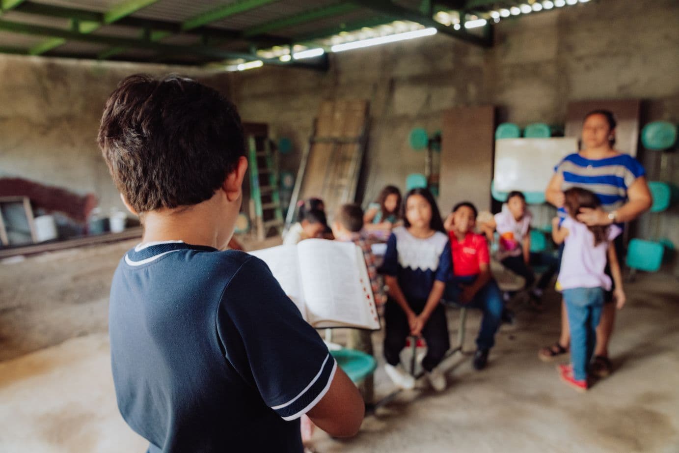 Poika lukee Raamattua, taustalla lapsia istumassa.