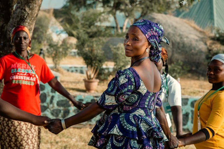 Naisia käsi kädessä piirimuodostelmassa ulkona. Etualalla nainen värikkäässä violetissa afrikkalaisessa mekossa ja huivissa kääntyy kohti kameraa.