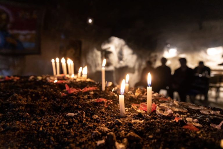 Hiekassa pystyssä olevia kynttilöitä palamassa, taustalla istuu tummia ihmishahmoja hämärän kirkon penkissä.