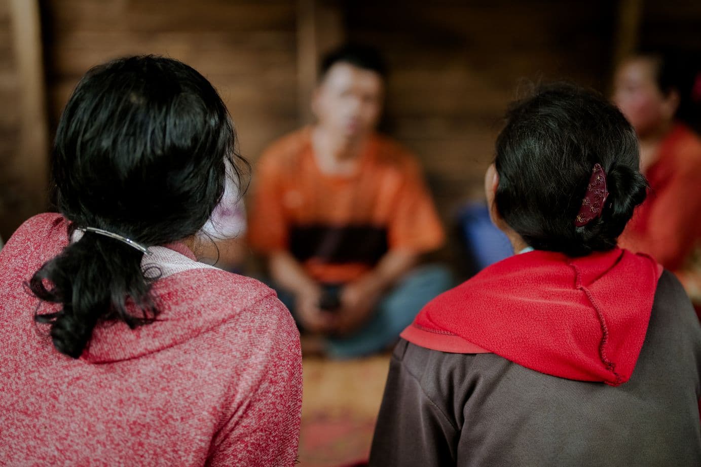 Kaksi nuorta istuu lattialla selin kameraan, heidän edessään, kasvot kameraan päin, istuu lattialla mies, jonka kasvot on sumennettu.