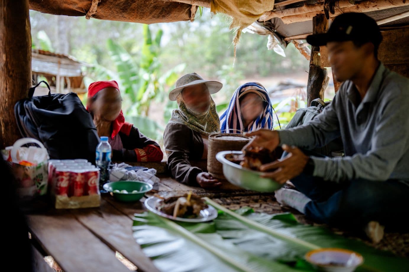 Mies istuu rakennuksen lattialla ja valmistaa ruokaa, taustalla kolme muuta ihmisitä sekä viidakkoa.