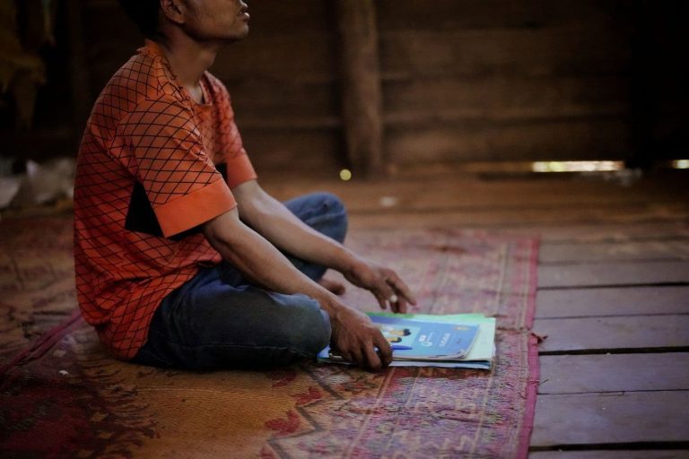 Mies istuu matolla lattialla edessään kasa koulukirjoja ja vihkoja.