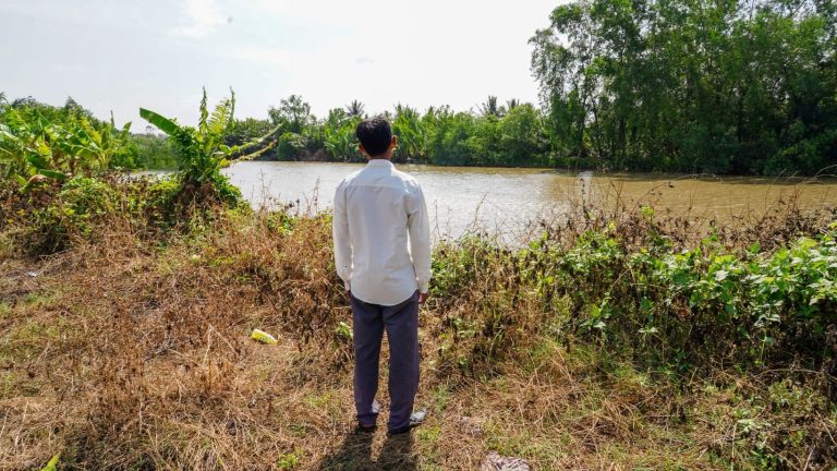 Mies seisoo joen rannalla selin kameraan, miehen takana joki ja joen takana metsää.