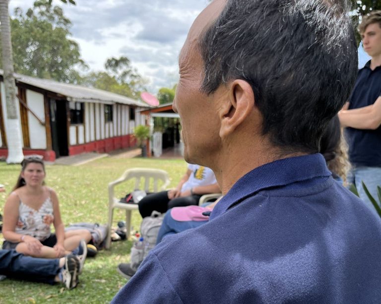 Mies kuvattuna takaviistosta, katsoo rakennusta, jonka edustalla nuoria istumassa maassa ja tuoleilla.