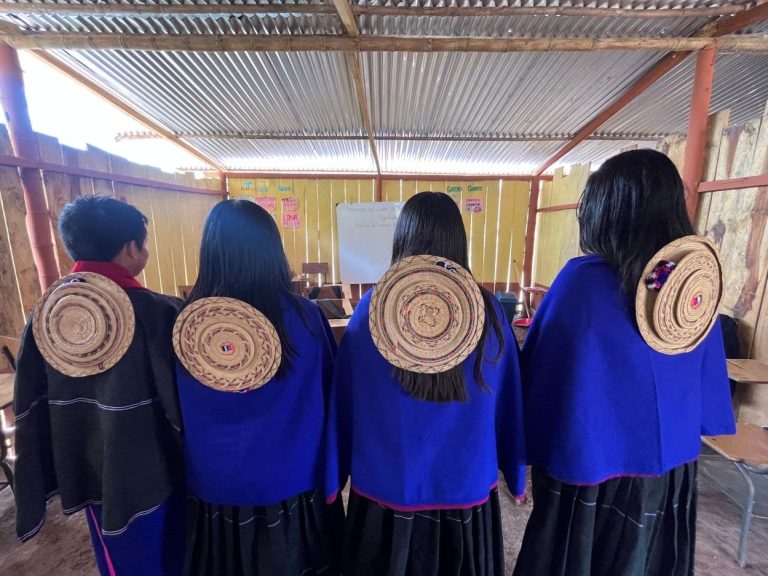 Neljä nuorta rivissä selin katsojaan, olkihatut niskassaan koululuokassa. Heidän edessään on pulpetteja ja valkotaulu.