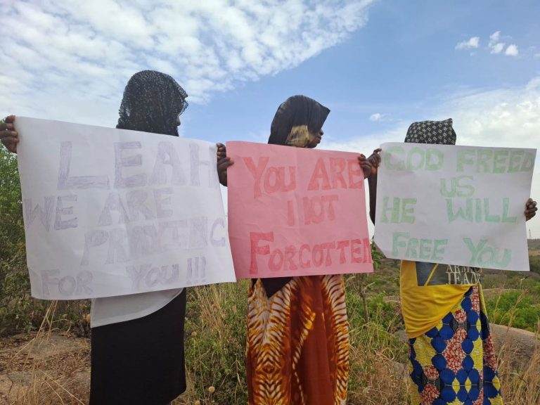 Kolme afrikkalaista nuorta naista käsissään suuret julisteet, joihin on kirjoitettu englanniksi tekstejä, joissa luvataan rukoilla Leah Sharibun puolesta.